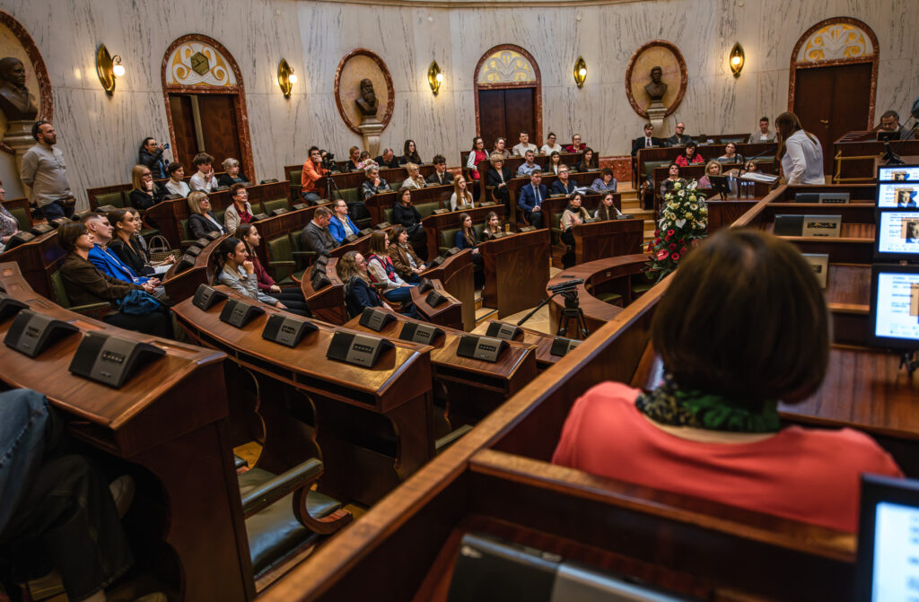 Tłum ludzi siedzących w sali Sejmu Śląskiego słucha przemówienia młodej dziewczyny